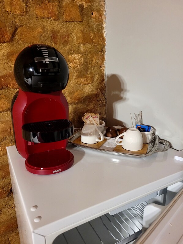 Une machine à café rouge est posée sur un comptoir blanc, et une soucoupe contient une tasse blanche, une carafe en verre et quelques dosettes de café.