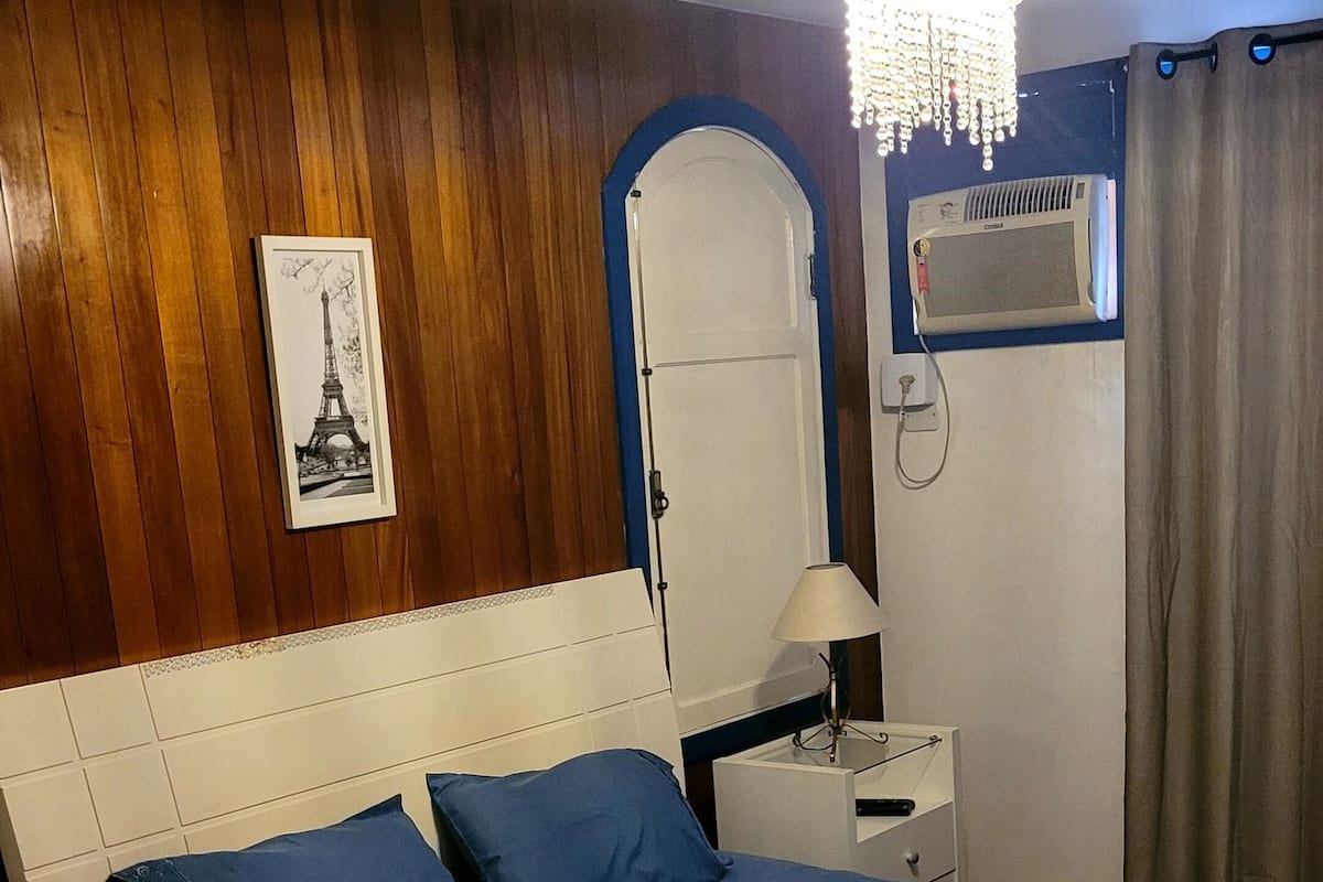 A bedroom with a bed, a nightstand, a lamp, a framed picture of the Eiffel Tower, and a window with curtains.