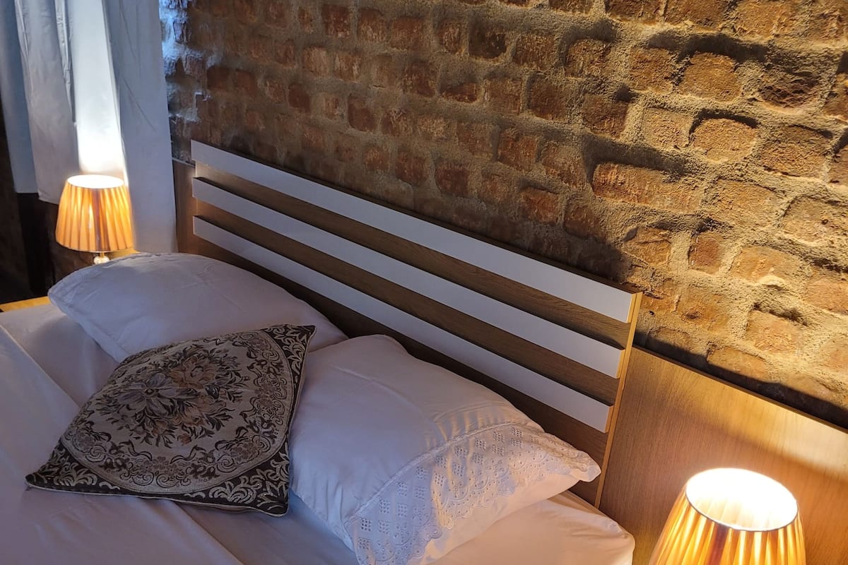 A neatly made bed with white linens and a patterned pillow, a wooden headboard, and a nightstand with a lamp and a laptop.