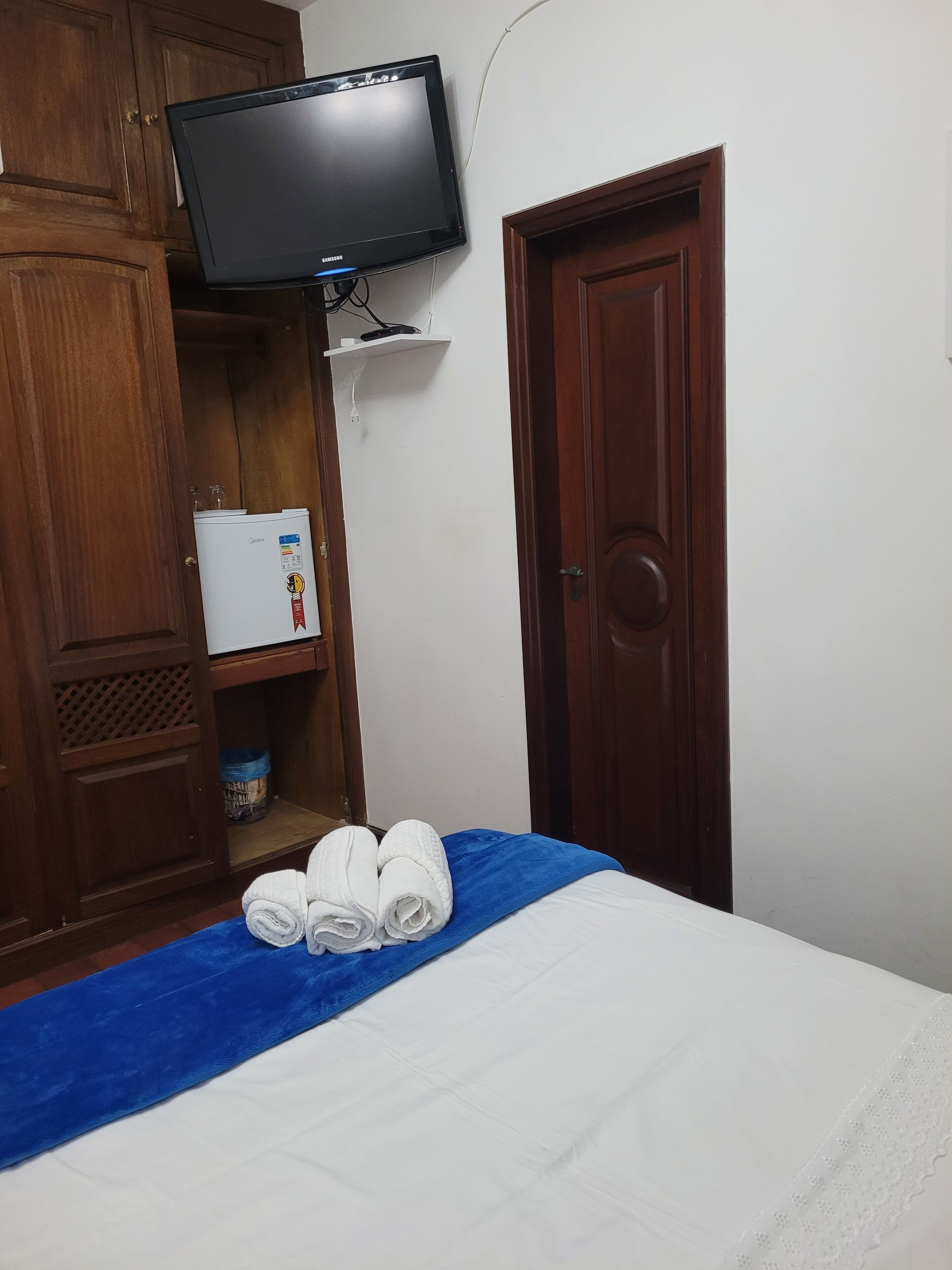 Quarto de hotel com uma cama, televisão fixada na parede, minibar e guarda-roupa de madeira.