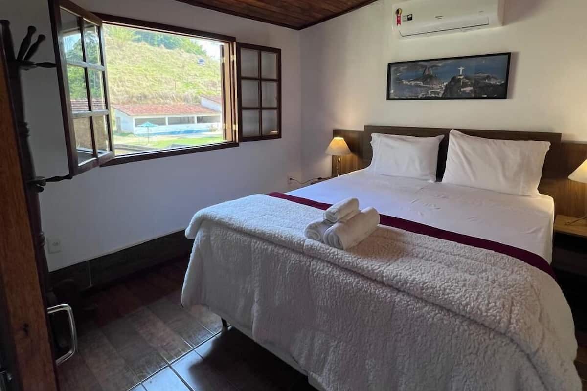 A bedroom with a wooden ceiling, a bed with white bedding, a window with a view of greenery, an air conditioner, and a painting on the wall.