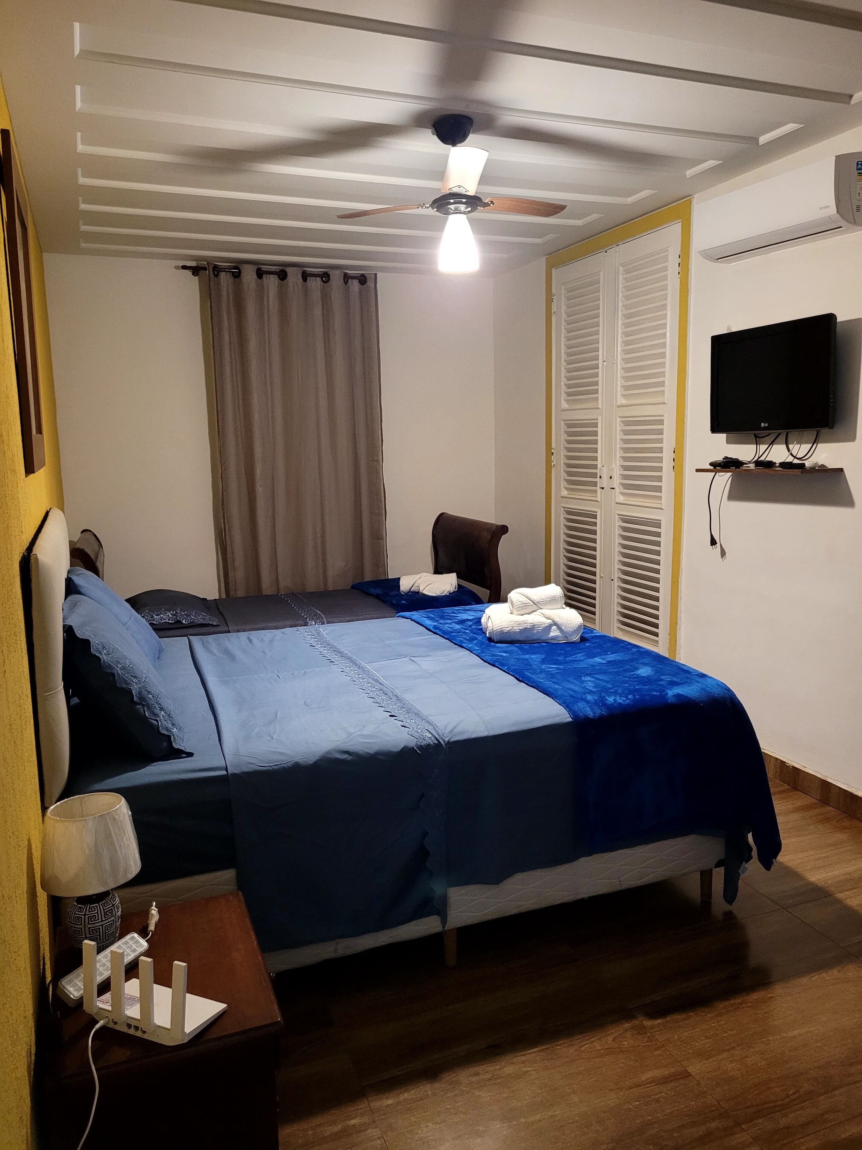 Una cama bien hecha con ropa de cama azul, un ventilador de techo, un televisor sobre un mueble bajo y una mesita de noche de madera con un teléfono y cargador.