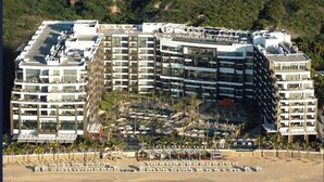 Un hotel de varios pisos con balcones, una playa en primer plano y montañas al fondo.