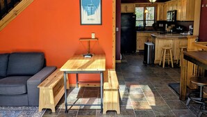 Un espacio de sala acogedor con un banco de madera, una mesita y un sofá. Se ve una cocina con gabinetes de madera y un refrigerador al fondo.