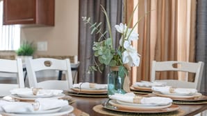 Una mesa de comedor dispuesta con platos blancos, cuencos con detalles dorados y caminos de mesa tejidos. En el centro, un jarrón azul rústico con flores blancas y follaje.