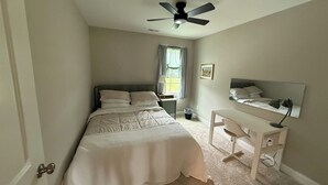 Une chambre à coucher comprenant un lit, un bureau, une chaise, un ventilateur de plafond, une fenêtre avec des rideaux et un miroir fixé au mur.