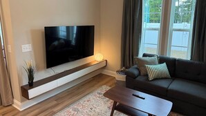 Un salon moderne avec un téléviseur à écran plat fixé sur une console en bois, un canapé de couleur foncée agrémenté de coussins et un tapis à motifs sur le plancher en bois.