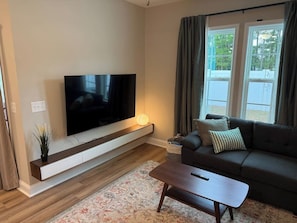 A modern living room with a flat-screen TV mounted on a wooden console, a dark-colored sofa with cushions, and a patterned rug on the wooden floor.