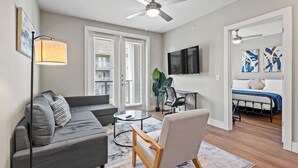 A modern living room with a grey sofa, a round coffee table, and a wooden chair. There is a ceiling fan, a TV mounted on the wall, and a bed in the adjacent room. - Comfy King bed | Pool + Gym + Parking + Stockyards (Fort Worth)