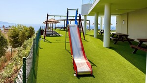 Un parque infantil con tobogán, columpios y mesas de picnic, con vistas panorámicas.