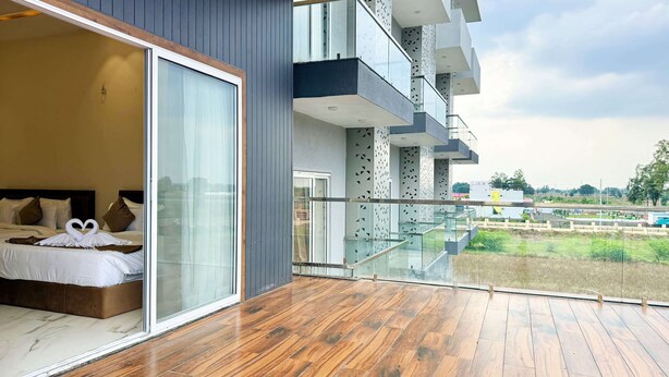 A modern hotel room with a large sliding glass door leading to a balcony. The room features a wooden floor, a bed with white bedding, and a heart-shaped towel decoration. The balcony has glass railings and offers a view of the surrounding landscape. - The Grand Omkara Hotel & Resorts (Khandwa)
