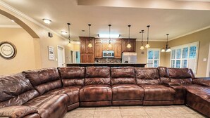 A spacious living room with a large leather sectional sofa, a kitchen area with wooden cabinets and a microwave, and a dining area with a table and chairs. - Spacious 3BR Home • Sleeps 6 • Open Living • Private Yard (Victoria)
