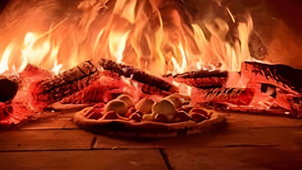 Ein Pizzaofen mit Feuer im Inneren, Holzscheiten auf dem Boden und einem Pizzastein davor.