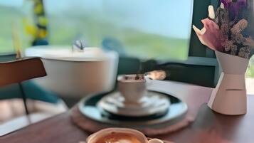 Una taza de café sobre un platito, un plato debajo, un jarrón con flores y una ventana con vistas a follaje.