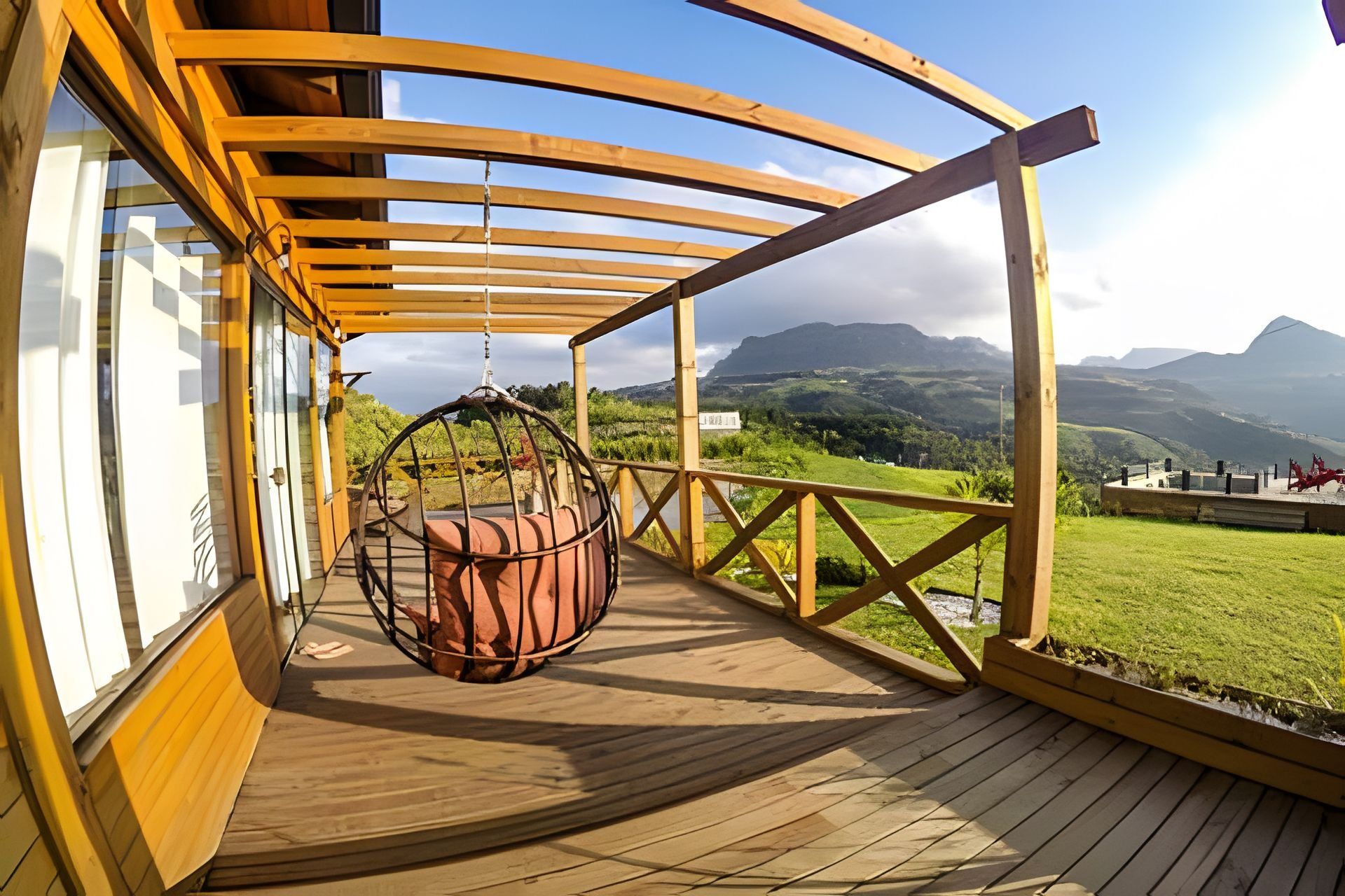 Una terraza de madera con una silla colgante, vistas a colinas y montañas verdes, y un cielo despejado.