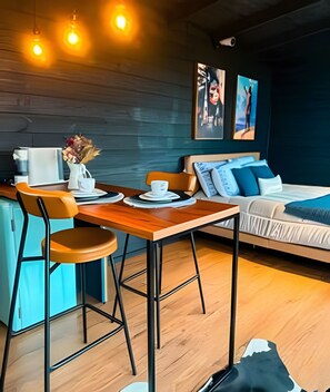 A modern room with a wooden dining table, two chairs, a sofa, and a bed. There are wall-mounted lights, a security camera, and artwork on the walls. - Refugio mirante da montanha (Alfredo Wagner)