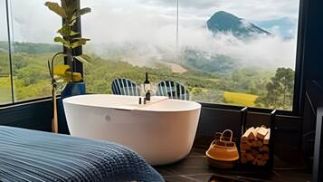 Ein Schlafzimmer mit großem Fenster, einem Bett, einer Badewanne und Blick auf eine gebirgige Landschaft.