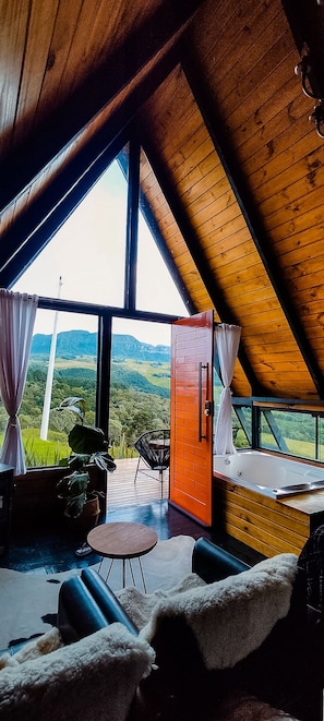 Um quarto com teto inclinado, grandes janelas, piso de madeira, um sofá, uma cadeira, uma pequena mesa e vista para uma paisagem verde.