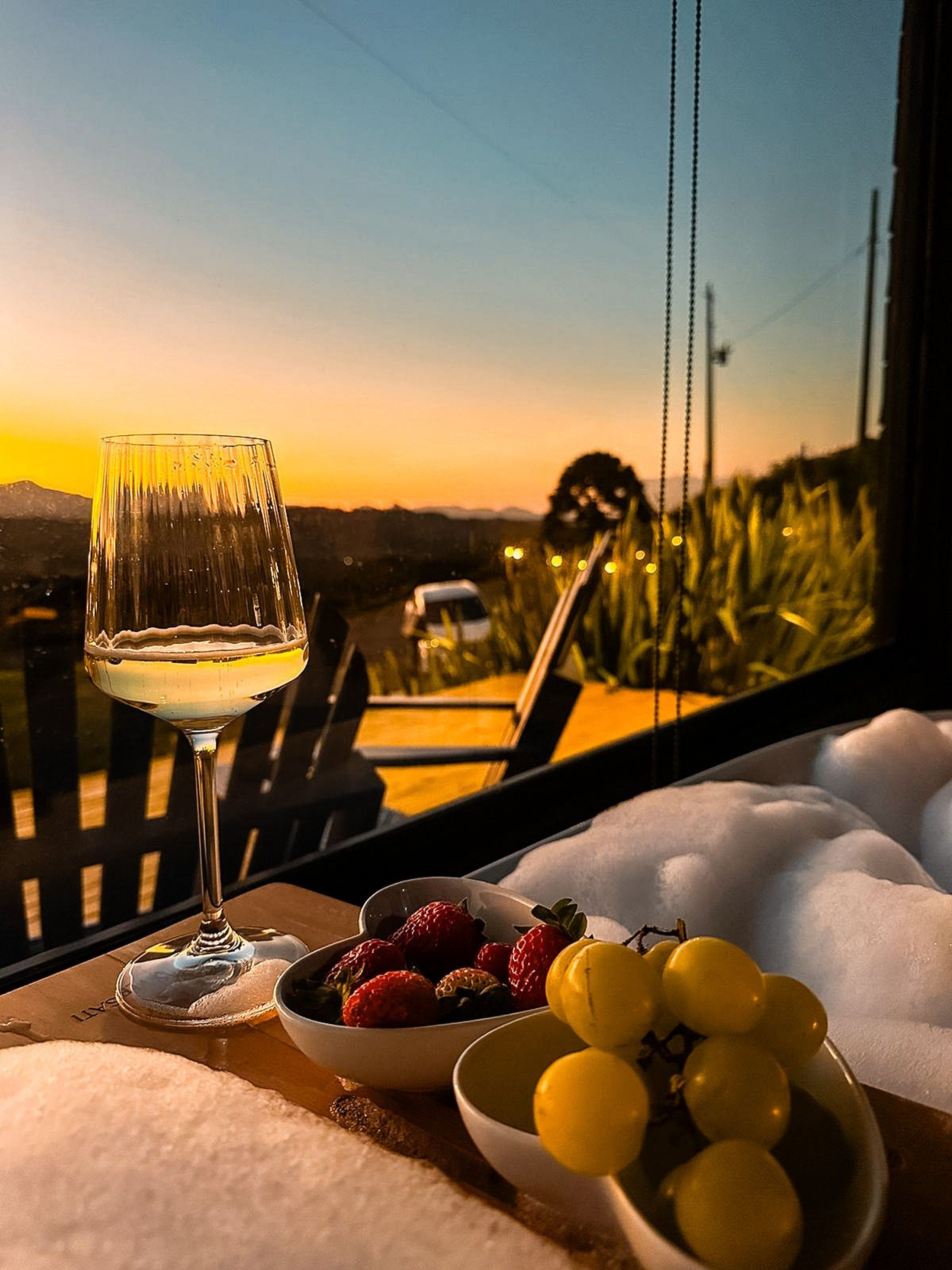 A table with a glass of white wine, a bowl of strawberries, and a bowl of yellow grapes, with a view of a sunset and mountains in the background.