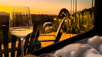 A table with a glass of white wine, a bowl of strawberries, and a bowl of yellow grapes, with a view of a sunset and mountains in the background.