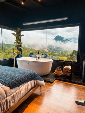 A bedroom with a large window, a bathtub, a bed, and a plant. - Refugio mirante da montanha (Alfredo Wagner)