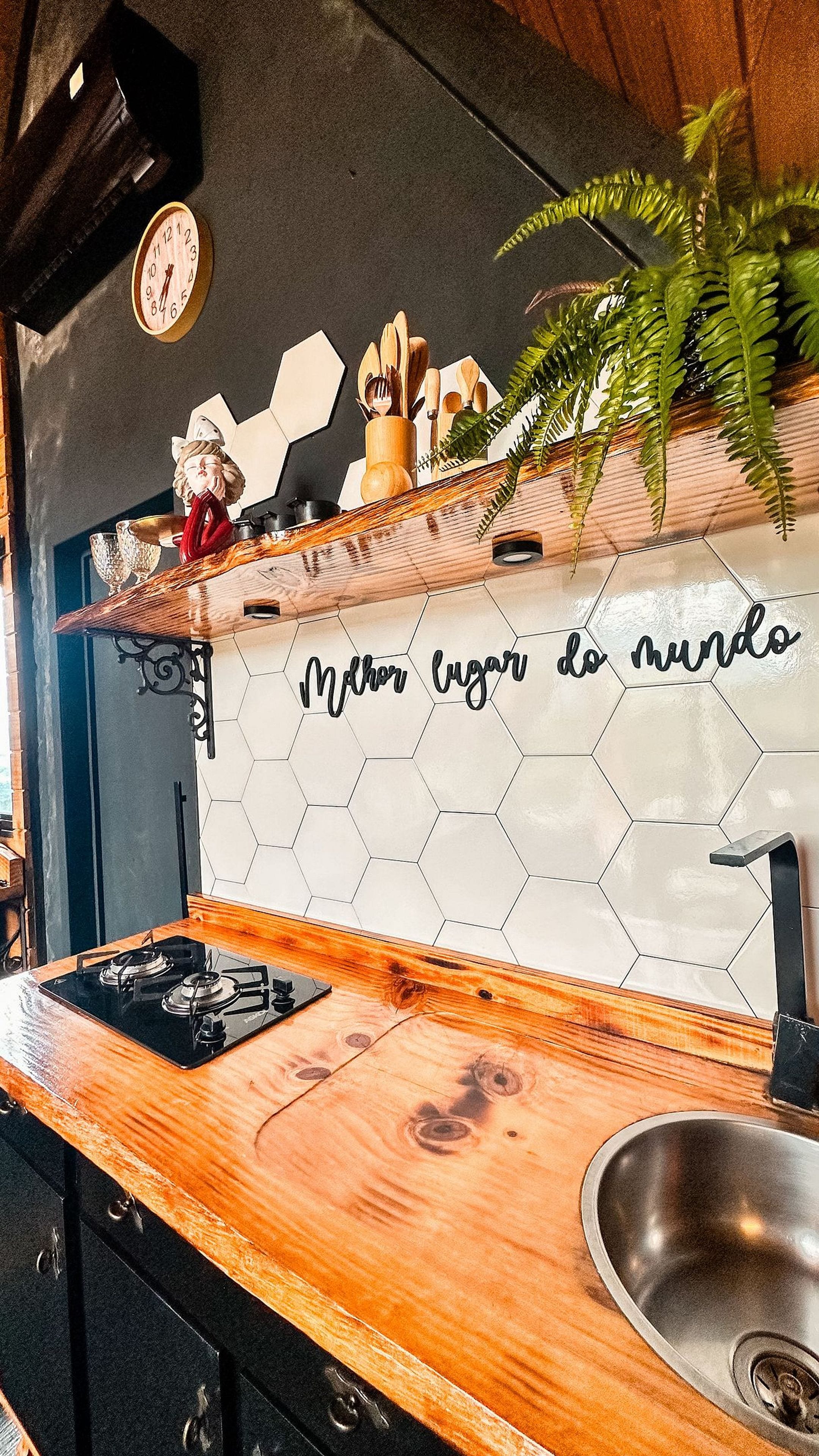 A kitchen with a wooden countertop, a stainless steel sink, a hexagonal tile backsplash with a phrase, and a plant.