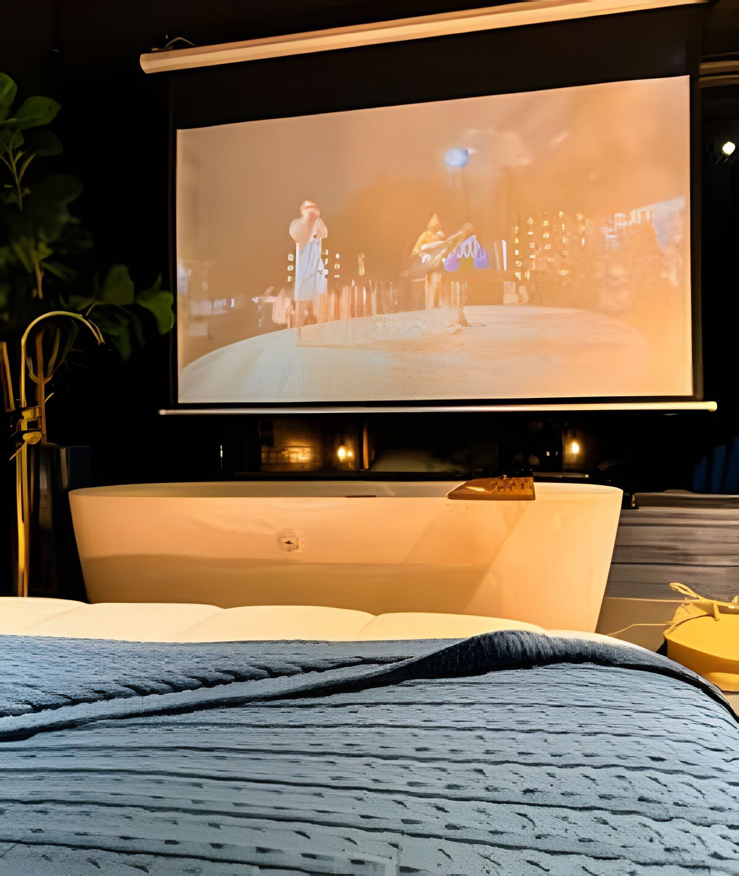 Ein Bett mit großem, darüber angebrachtem Fernseher, einem Nachttisch und einer Pflanze im Hintergrund.