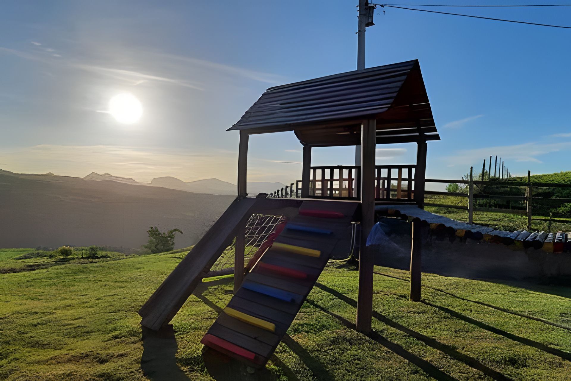 Um parquinho com um escorrega e um pequeno abrigo, com montanhas e um céu claro ao fundo.