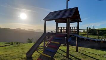 Um parquinho com um escorrega e um pequeno abrigo, com montanhas e um céu claro ao fundo.