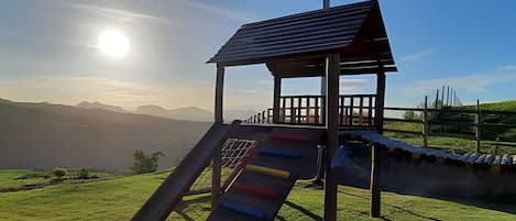 A playground with a slide and a small shelter, set against a backdrop of mountains and a clear sky.