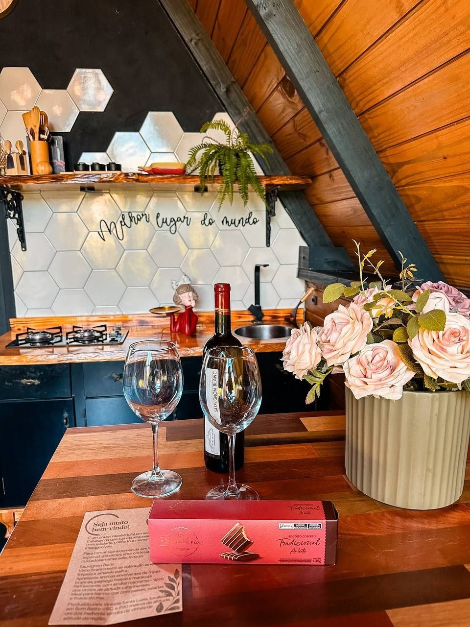 A kitchen with a hexagonal tile backsplash, a wooden countertop, a bottle of wine, two wine glasses, a vase of flowers, and a menu on the table.