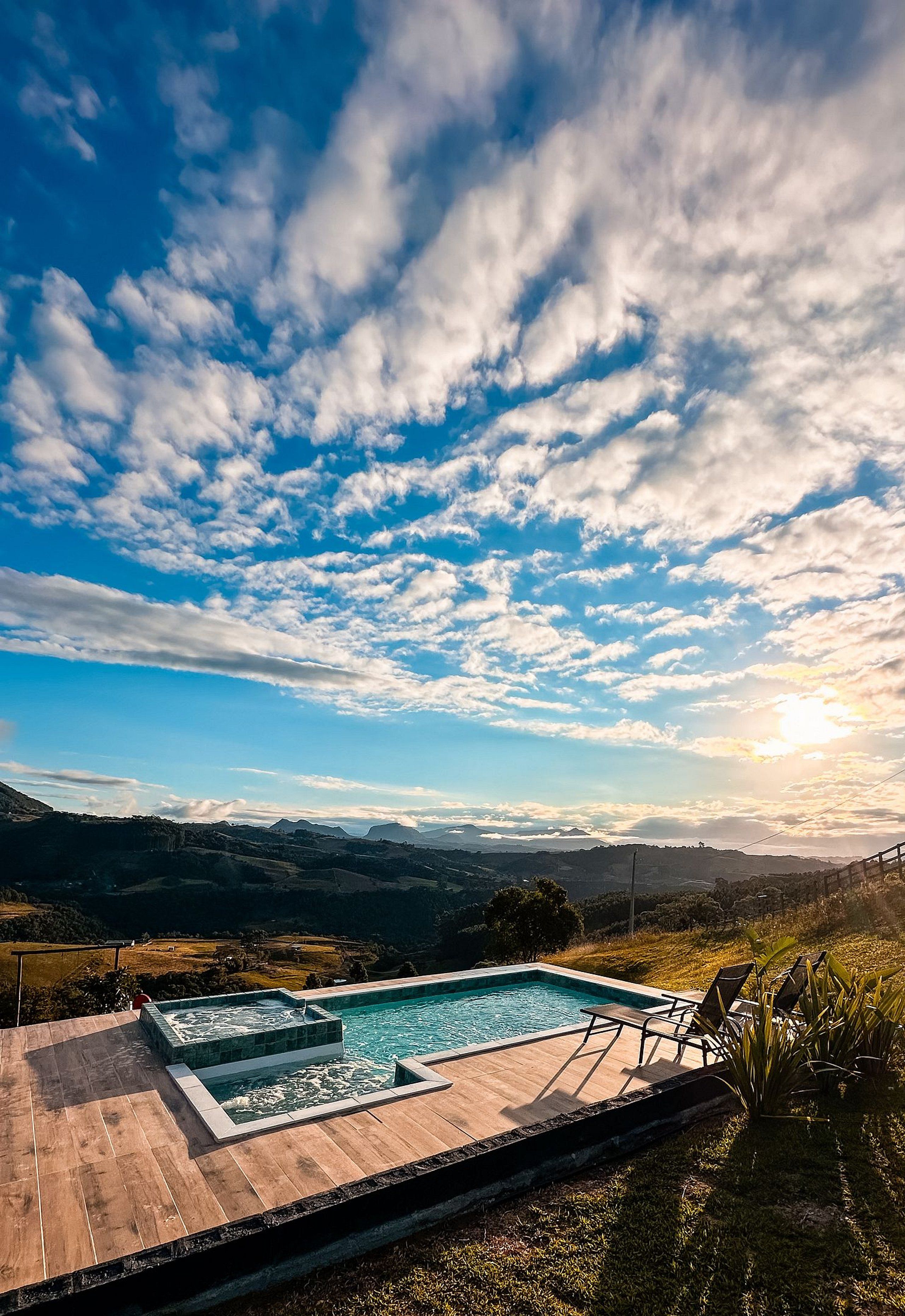 Una piscina con cubierta de madera, rodeada de césped y plantas, con vistas a colinas distantes y un cielo parcialmente nublado.