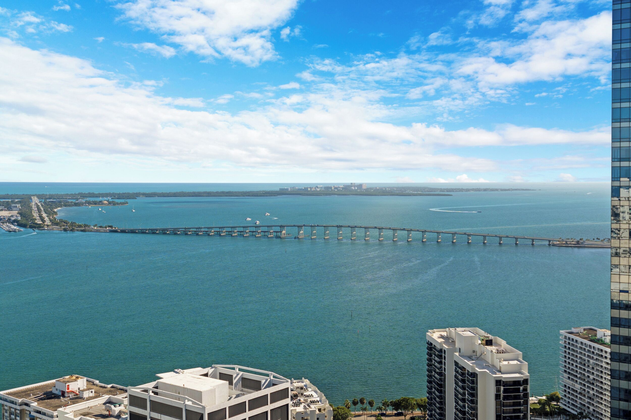 A coastal cityscape with a prominent bridge extending into the water, modern buildings, and a clear sky.
