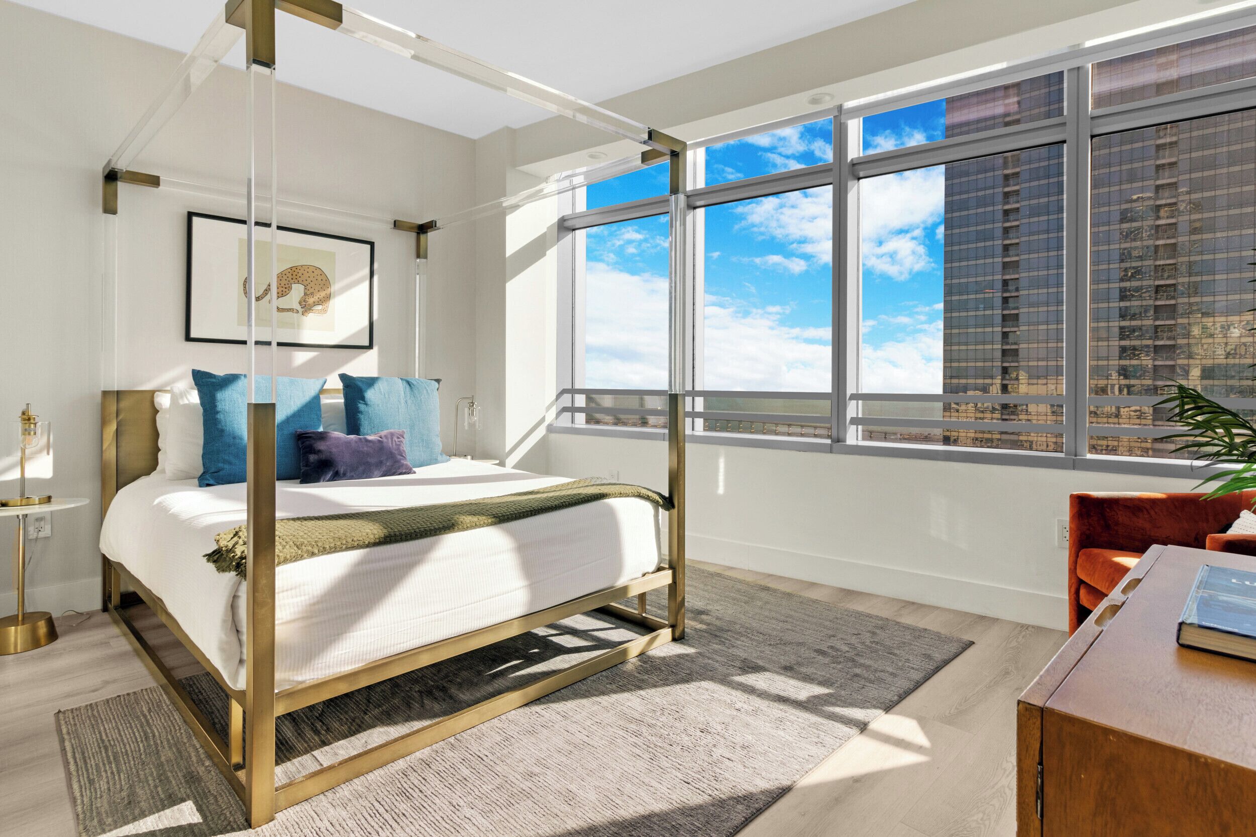 A modern bedroom with a four-poster bed, a large window with a city view, and a small desk with a chair.