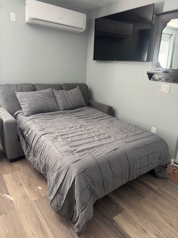 A small, modern bedroom with a grey sofa bed, a mounted TV, and an air conditioning unit.