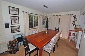 Une salle à manger avec une table en bois, des chaises et un lustre. Les murs sont ornés de tableaux encadrés et un petit sapin de Noël est installé dans la pièce.