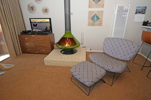 Ett mysigt vardagsrum med en vedeldad kamin, en tv på ett träskåp och mönstrade kuddar i en fåtölj.