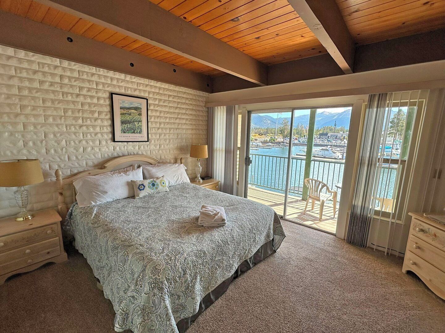 A bedroom with a large bed, a wooden ceiling, a white brick wall, and a view of a marina through a sliding glass door.