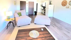 Habitación de hotel compacta con dos camas, una mesita, una lámpara y una kitchenette al fondo.