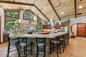 Una cocina amplia con una isla grande, vigas de techo de madera y ventanas panorámicas con vistas a árboles.