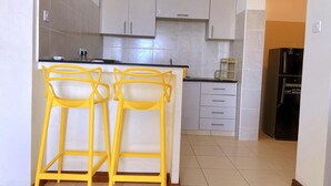 A kitchen with two yellow bar stools, a black refrigerator, and white cabinets. - Nova Stays 1-Bedroom Apartment with Private Pool & Uninterrupted Seaview (Mombasa)
