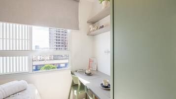 Een kleine, lichte kamer met een bed, een bureau met twee bekers, een raam met jaloezieën en uitzicht op gebouwen.