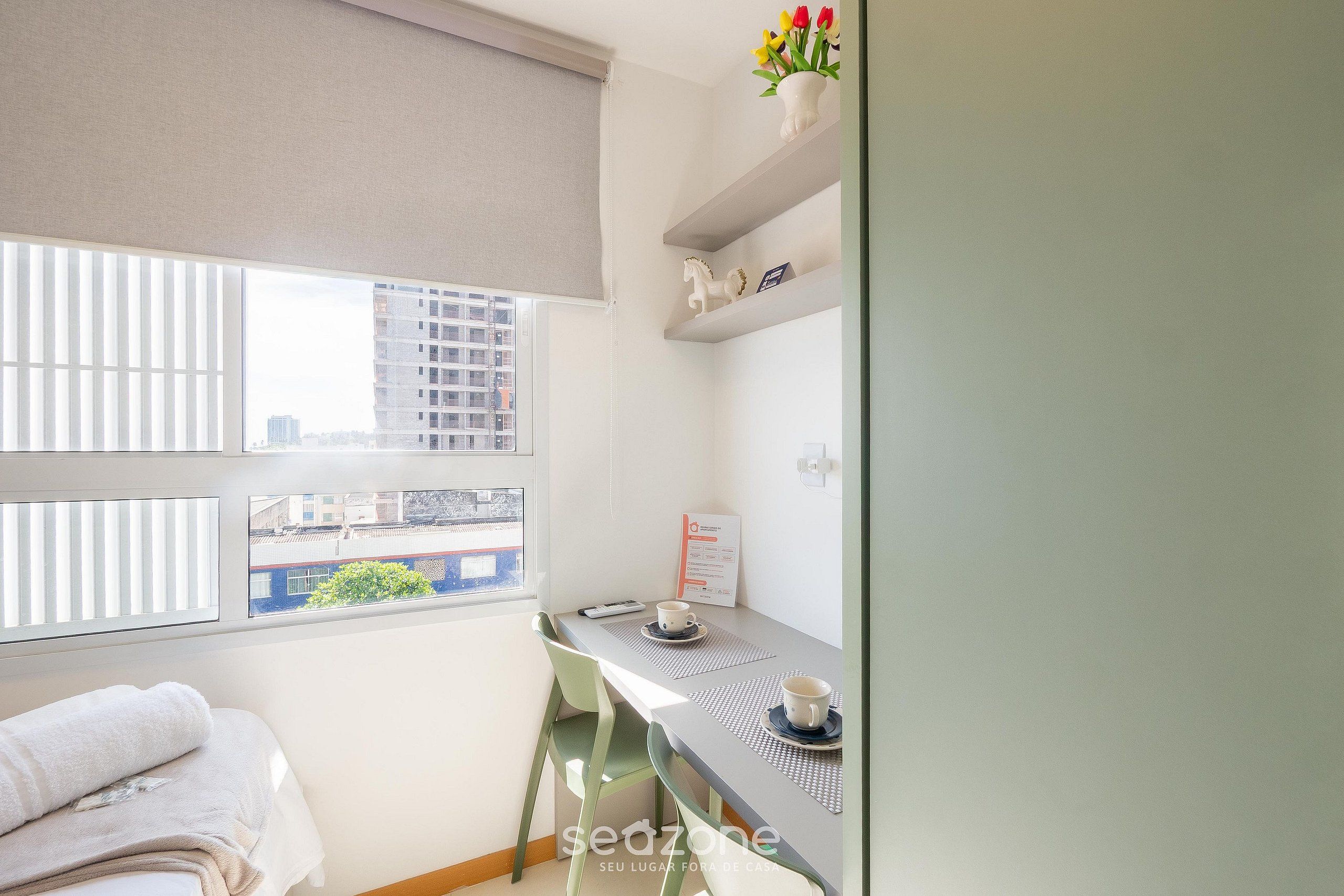 A small, bright room with a bed, a desk with two cups, a window with blinds, and a view of buildings.