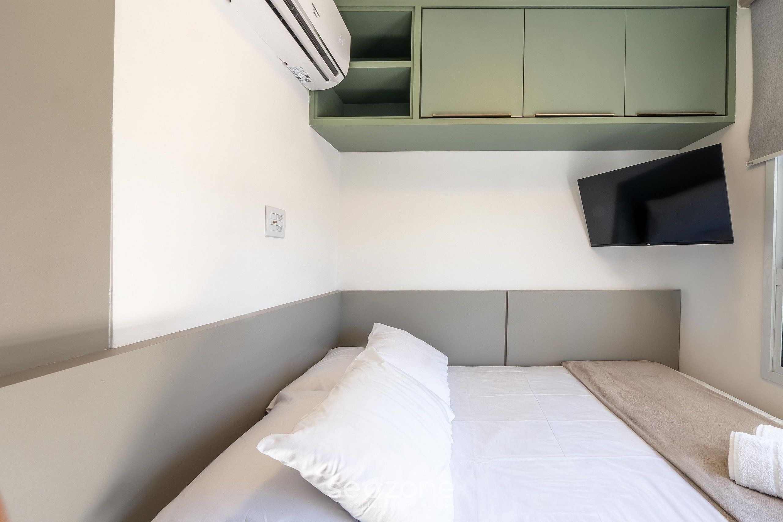 A small, neatly made bed with white bedding and a single pillow, a green cabinet above, and a flat-screen TV mounted on the wall.