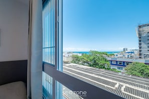 Blick aus einem Fenster auf ein Gebäude mit einer Metalldach, einem Balkon und einem wolkenlosen, blauen Himmel.