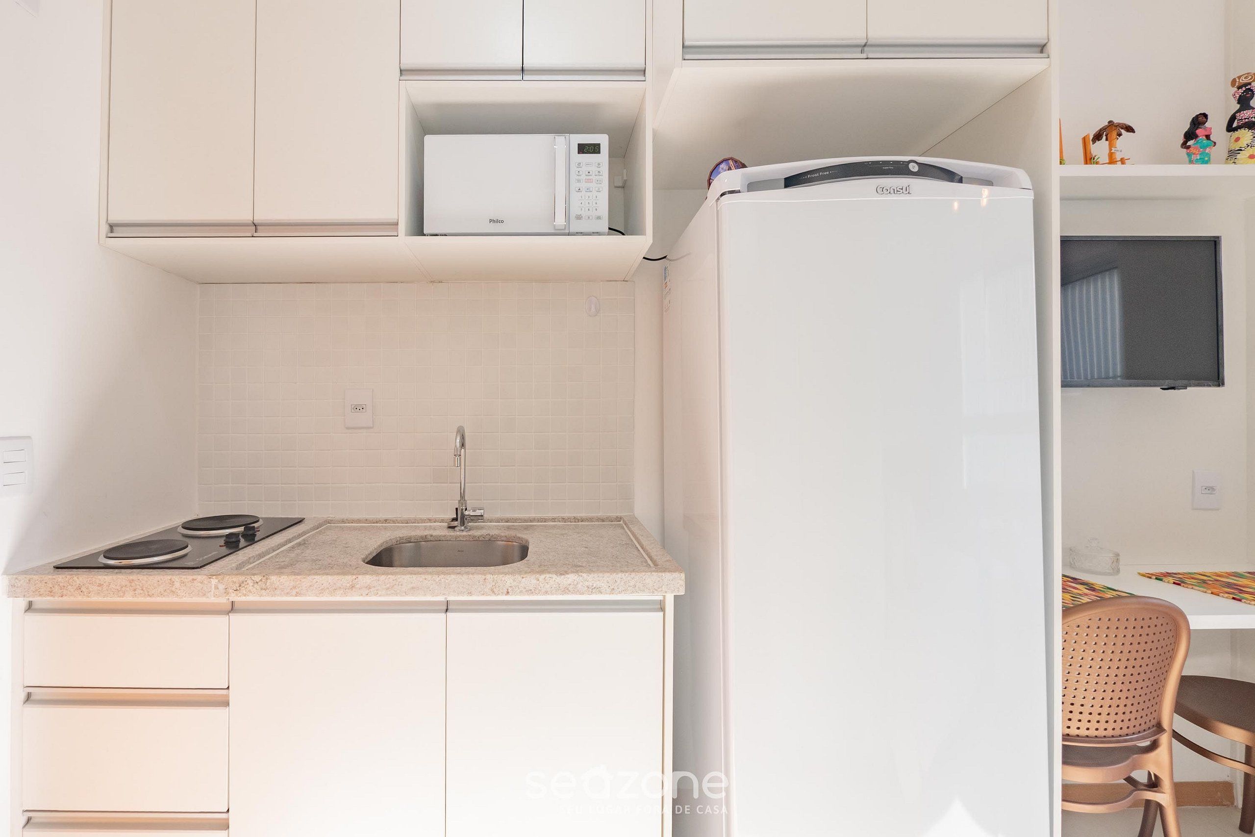 Cocina compacta con armarios blancos, microondas, refrigerador, pileta y un pequeño comedor con una silla.