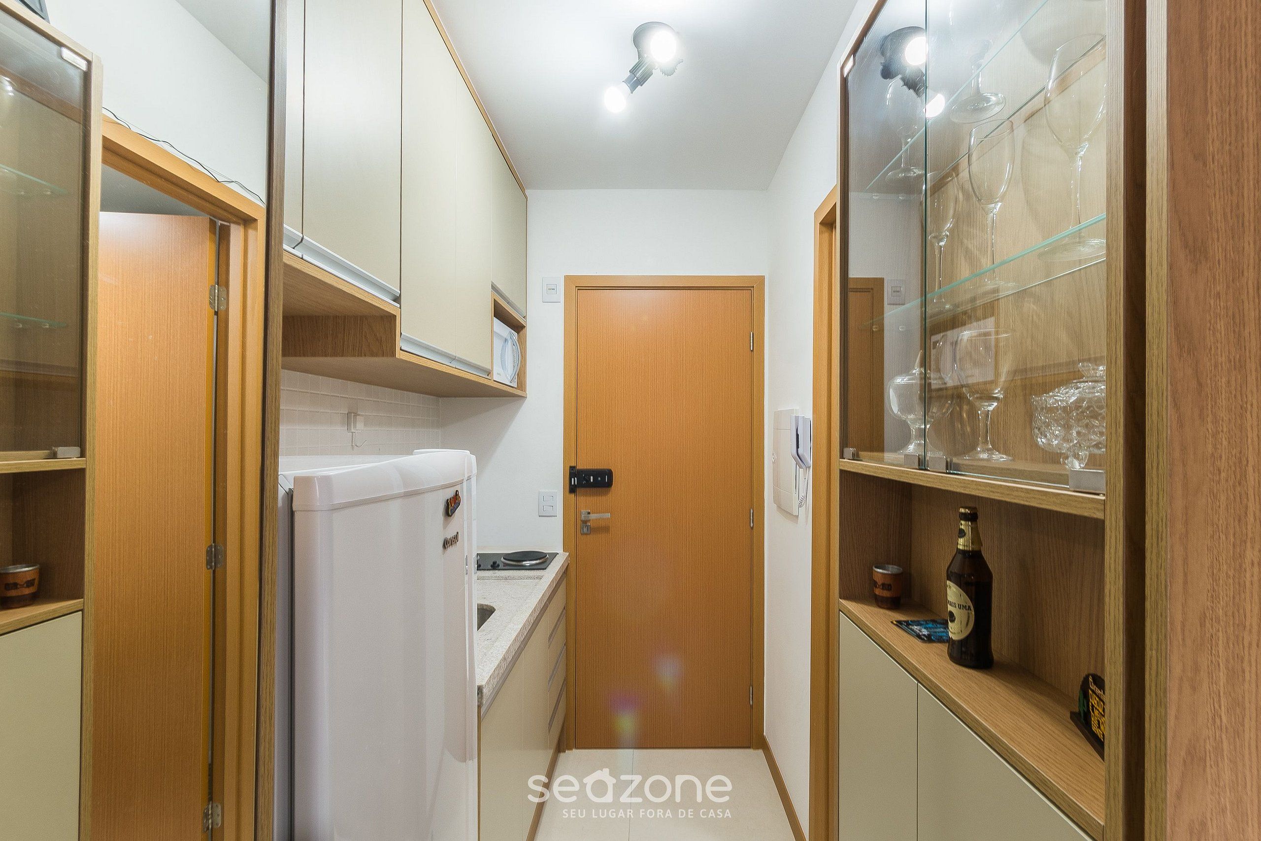 A narrow kitchen with wooden cabinets, a white refrigerator, and a glass display case with wine glasses.