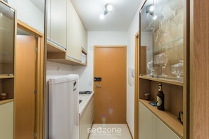Eine schmale Küche mit Holzschränken, einem weißen Kühlschrank und einer Glastheke mit Weingläsern.
