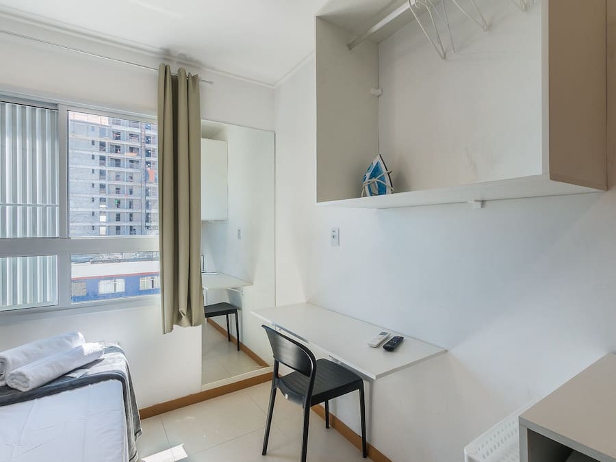 Een hotelkamer met een bed, een bureau, een stoel, een klein plankje en een raam met gordijnen.