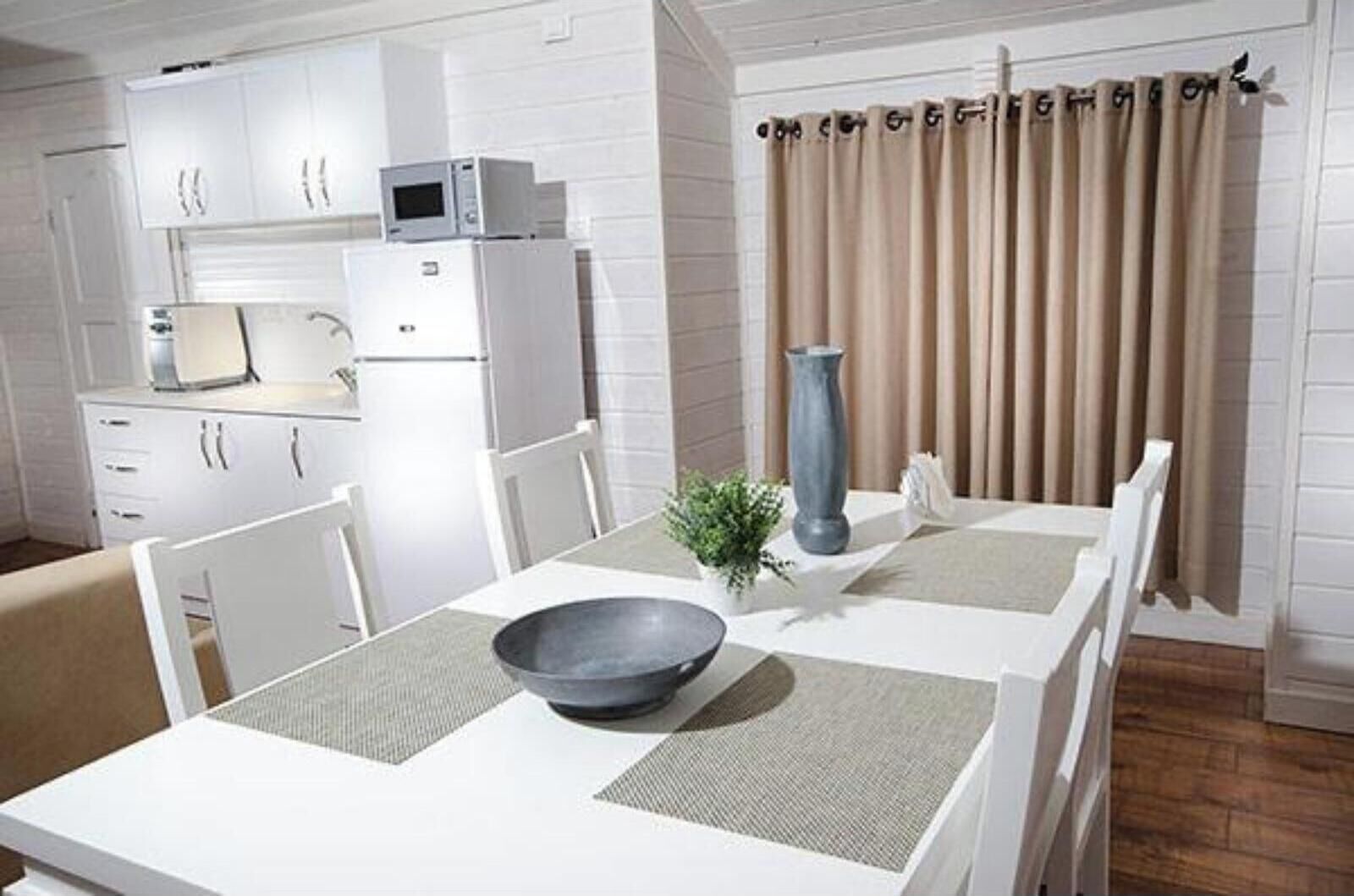 Een kleine keuken met witte kasten, een magnetron en een koelkast. Er staat een eettafel met een kom en een vaas erop.